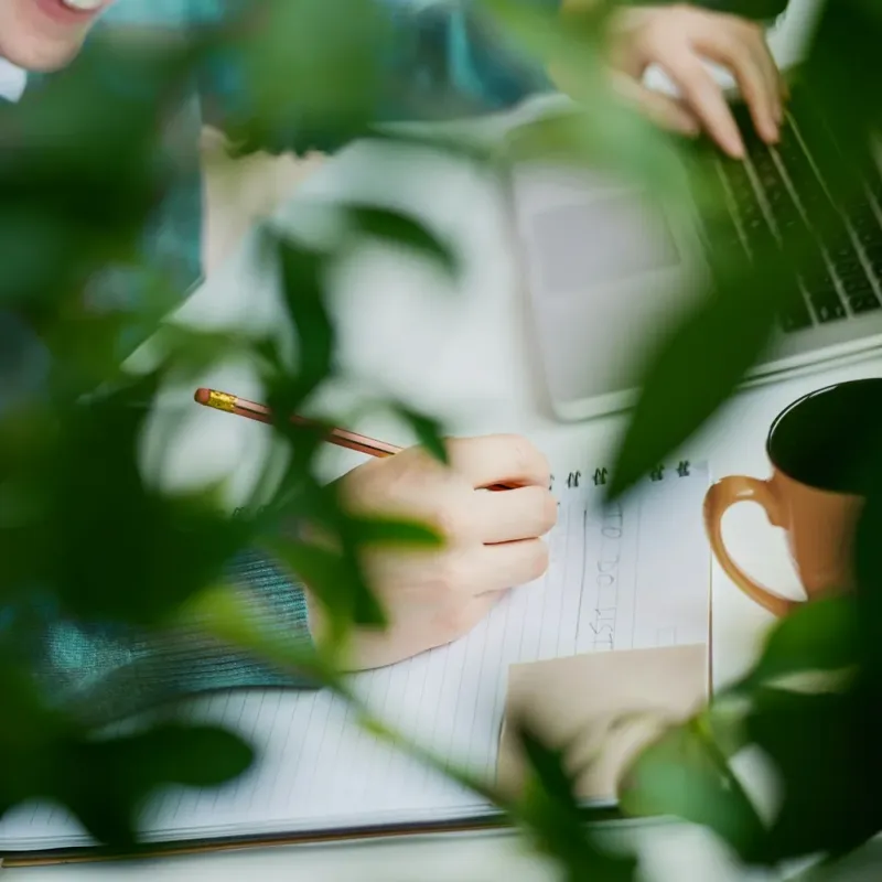Someone writing notes by hand beside a laptop, seen through leaves