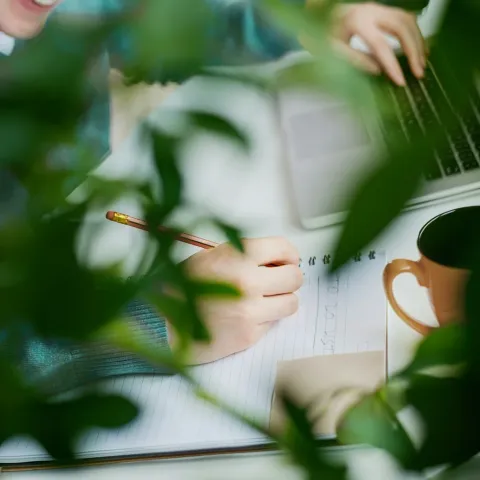 Someone writing notes by hand beside a laptop, seen through leaves