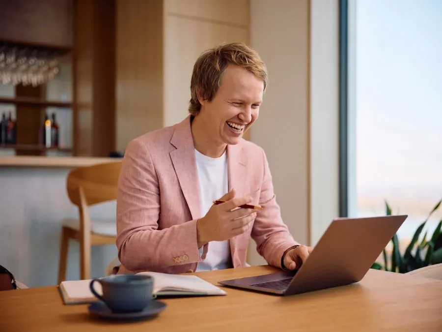 A business owner laughing during a video strategy call on his laptop, in a light-filled room with a window behind him